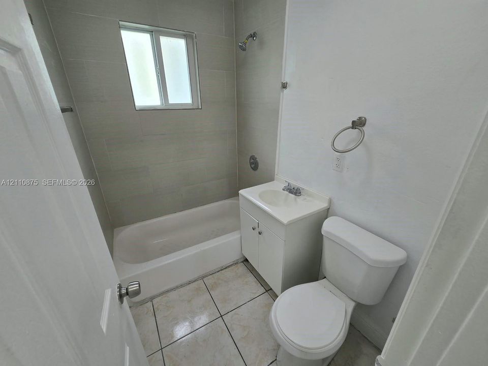 Bathroom featuring light-toned tile flooring, a bathtub with a shower head, wall-mounted shower tile surround, a white vanity cabinet with an integrated basin, and a wall-mounted ring towel holder