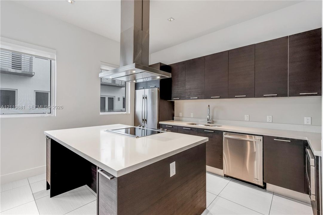 Kitchen featuring flat-panel dark wood cabinetry, a central island with a quartz countertop, stainless steel appliances, a ceiling-mounted stainless steel range hood, and tile flooring