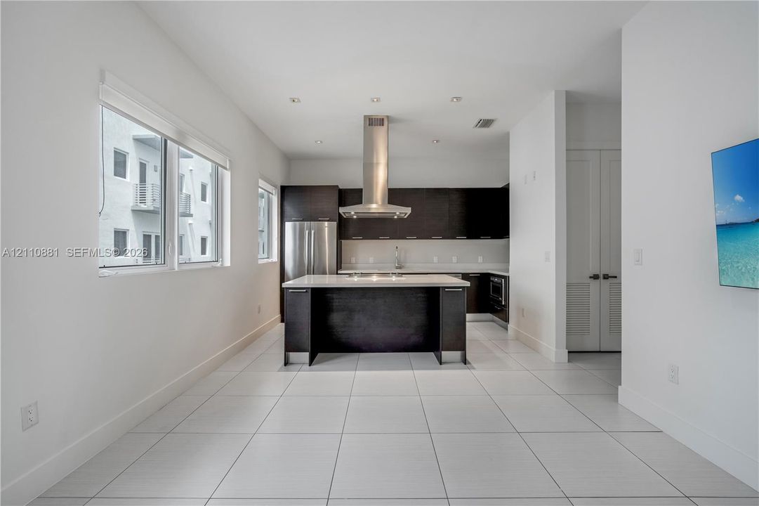 Modern kitchen featuring dark cabinetry, a central island with a light-toned countertop, stainless steel appliances, a ceiling-mounted range hood, and large-format tile flooring