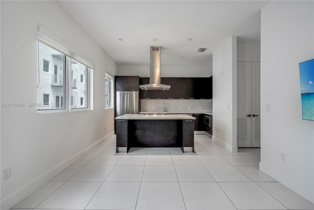 Kitchen featuring dark wood cabinetry, a central island with a stone-surface countertop, stainless steel appliances, a ceiling-mounted range hood, and large-format tile flooring