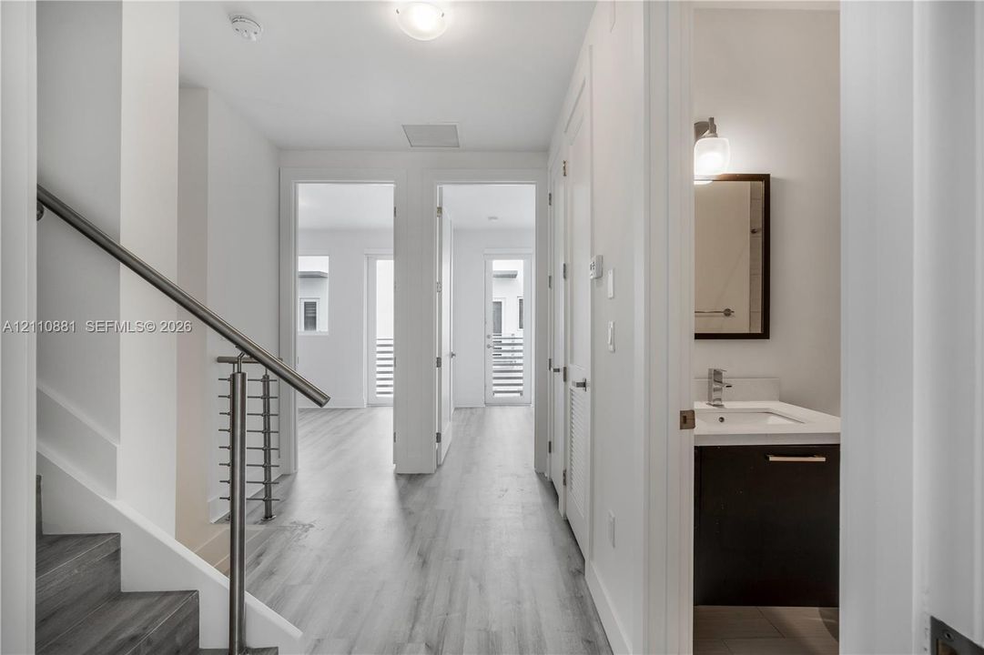 Interior hallway featuring wood-finish flooring, a staircase with metal railing, and a bathroom equipped with a dark wood-finish vanity and integrated sink