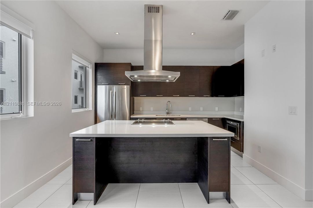 Modern kitchen featuring dark wood-finish cabinetry, a large center island with a stone-composite countertop, stainless steel appliances, and an overhead stainless steel vent hood