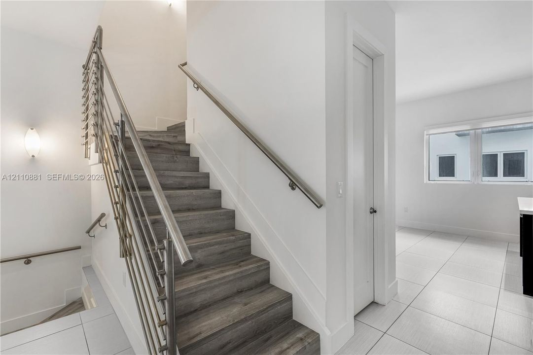 Staircase featuring wood-finish treads, metal horizontal railing, wall-mounted handrail, and large-format tile flooring
