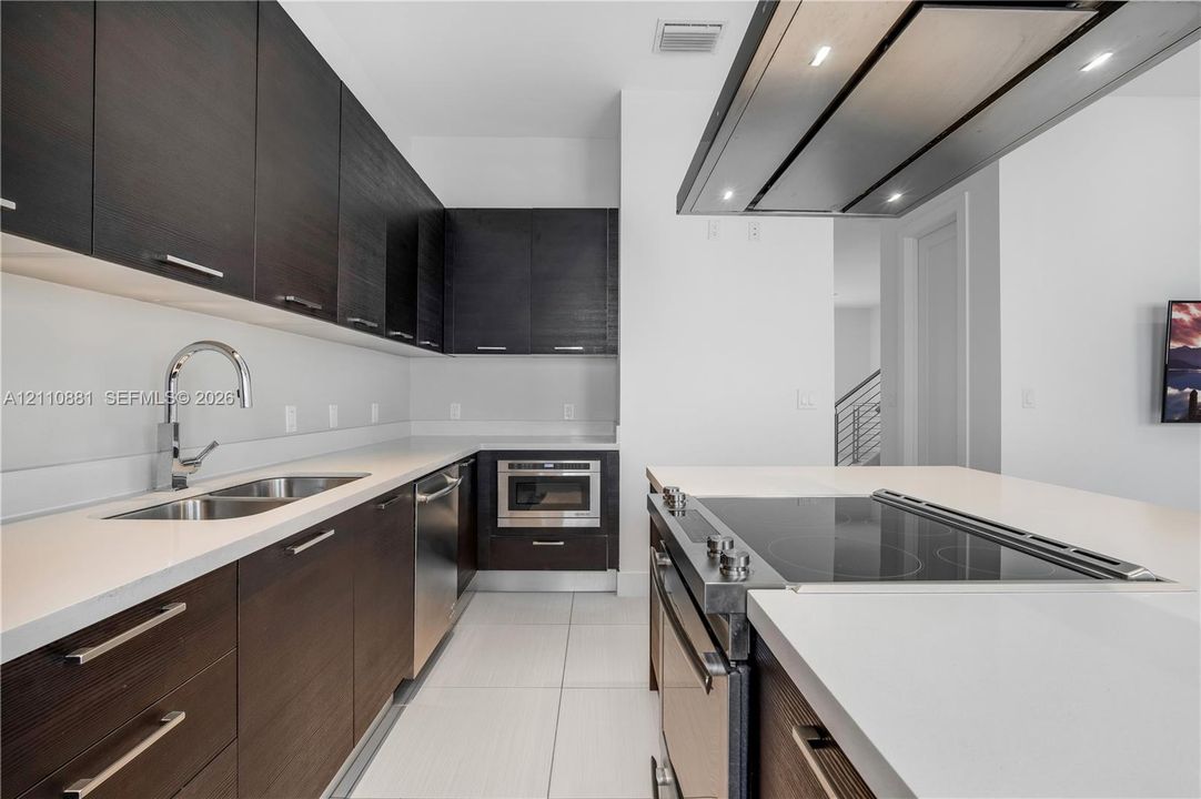 Kitchen featuring dark-finish flat-panel cabinetry, light-toned stone countertops, a large center island with an integrated cooktop, a stainless steel undermount sink, and a stainless steel range hood