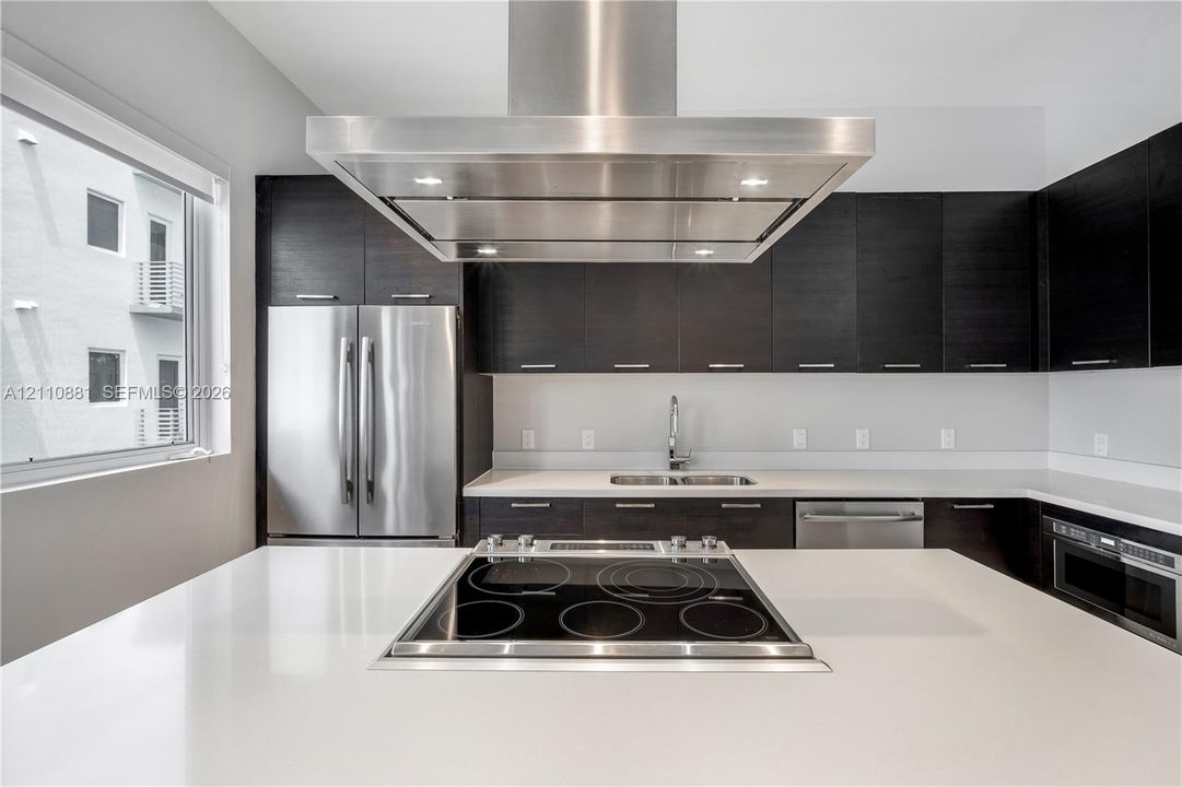 Kitchen featuring dark flat-panel cabinetry, light solid-surface countertops, a stainless steel island-mounted range hood, a built-in electric cooktop, and stainless steel appliances