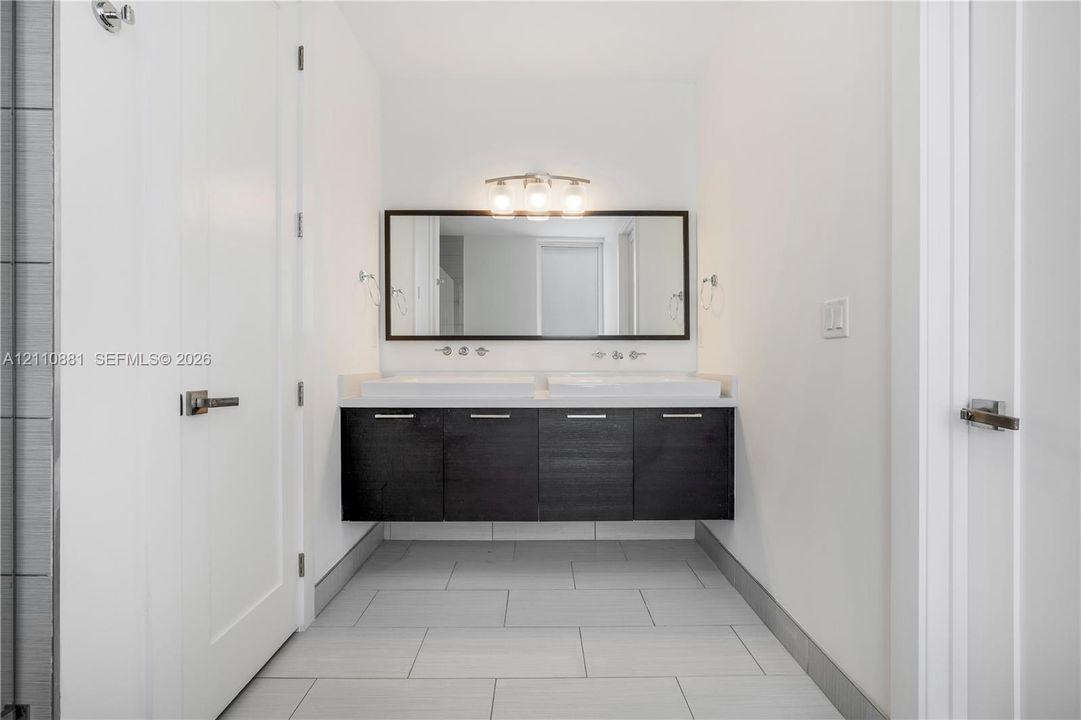 Floating dual-sink vanity with dark cabinetry, white countertop, rectangular vessel sinks, chrome wall-mounted faucets, and framed vanity mirror with integrated three-bulb fixture
