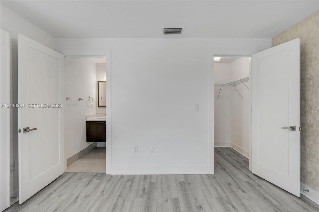 Wood-finish flooring, two white swing doors, a storage closet with built-in wire shelving, and an en-suite area featuring a dark wood-finish vanity with a rectangular sink