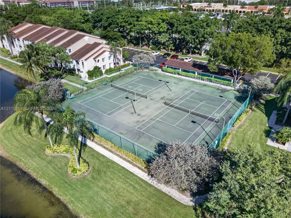 Fenced dual tennis courts featuring flood lighting, nestled within a landscaped community grounds with waterfront access and mature trees
