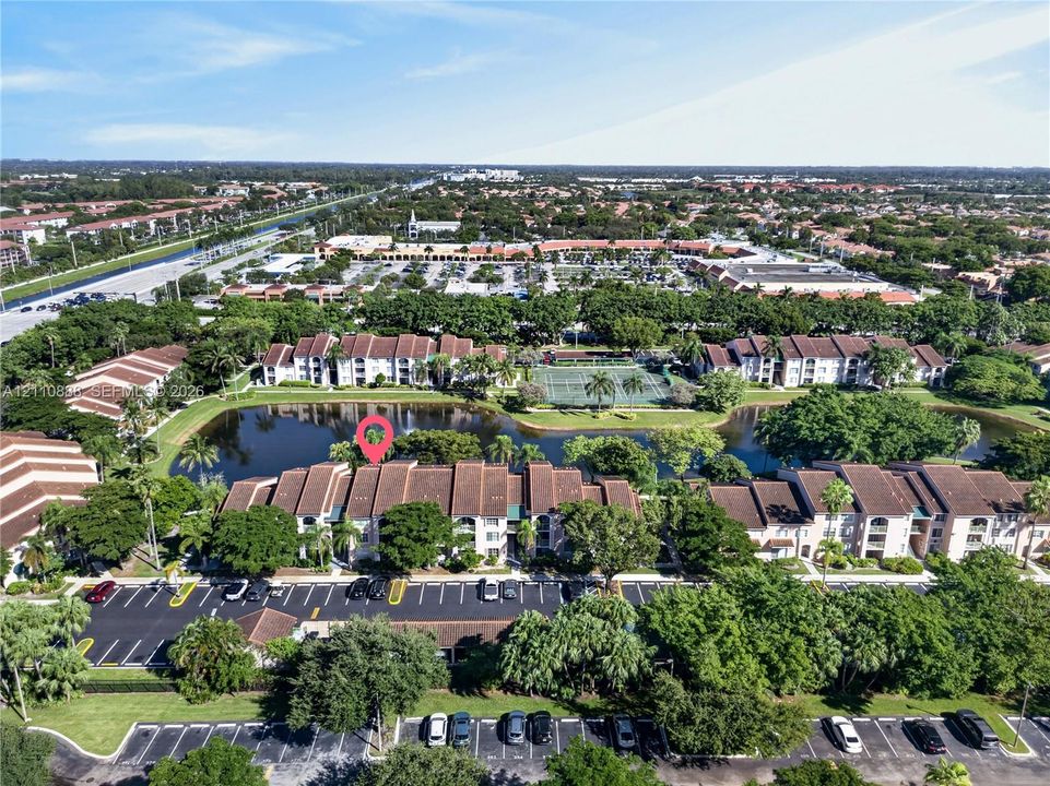 Aerial perspective of residential complex featuring a central pond, multi-story buildings with tile roofing, surface parking, and an on-site tennis court