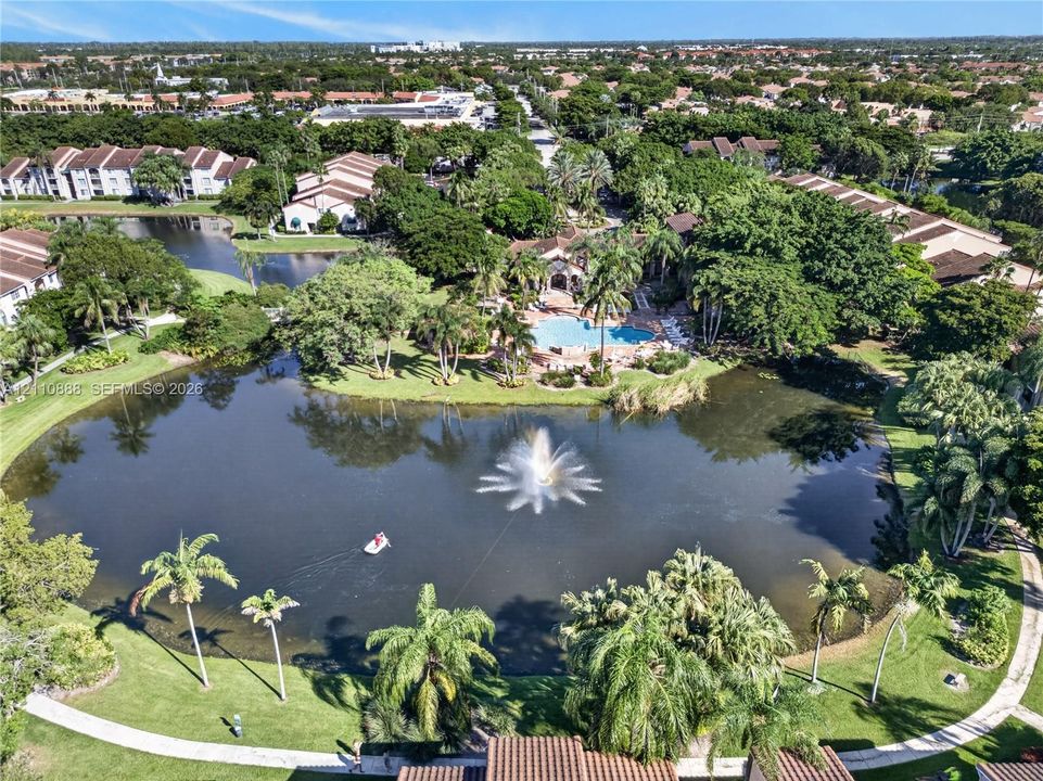 Aerial perspective of a lake featuring a central fountain, surrounding palm trees, paved walking paths, and a community swimming pool