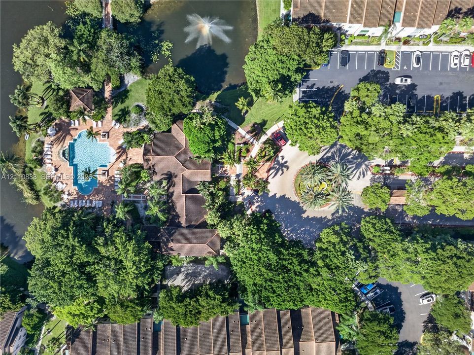 Aerial perspective of a community featuring a pool, a pond with a fountain, mature landscaping, and paved parking areas