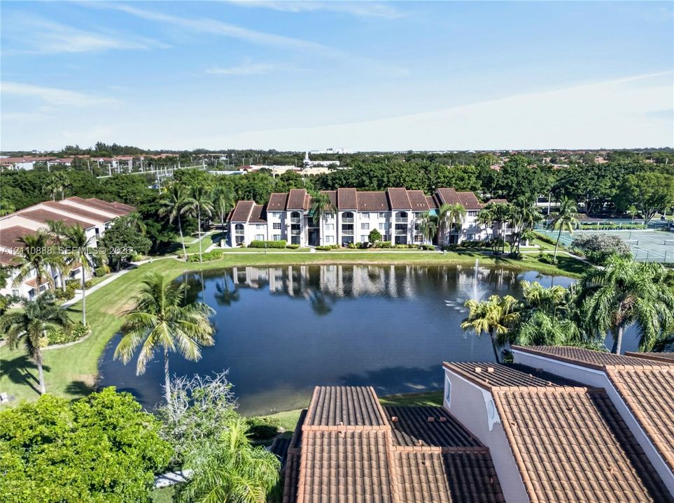 Aerial perspective featuring a multi-story residential building with a tile roof, situated beside a water feature with an integrated fountain, surrounded by mature palm trees and landscaped green space
