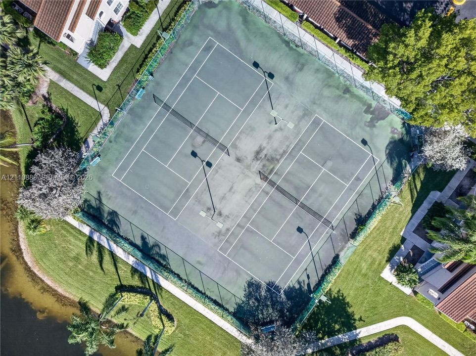 Dual tennis courts enclosed by chain-link fencing, featuring overhead lighting and perimeter landscaping