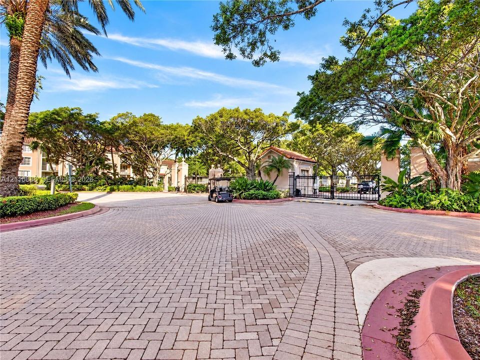 Gated community entrance featuring decorative paver driveway, mature landscaping, and established trees