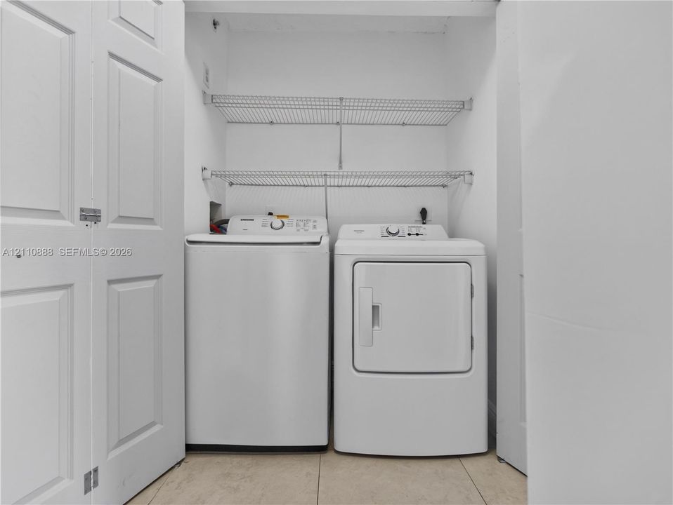 Laundry area featuring a top-load washer, a front-load dryer, dual wire shelving units, and tile flooring, enclosed by white bi-fold doors