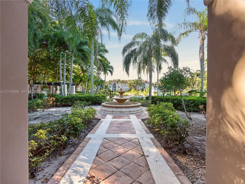 Outdoor courtyard featuring a central stone tiered fountain, decorative paver walkway with stone accents, manicured shrubbery, and mature palm trees