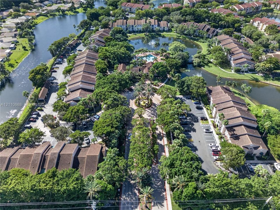 Aerial view of a community featuring waterfront surroundings, extensive mature landscaping, a central swimming pool, and paved surface parking