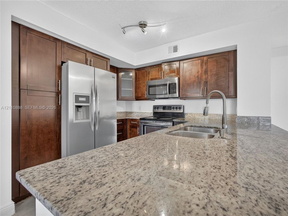 Kitchen featuring dark wood-finish cabinetry, stone countertops, stainless steel appliances, a dual-basin undermount sink with a gooseneck faucet, and track lighting