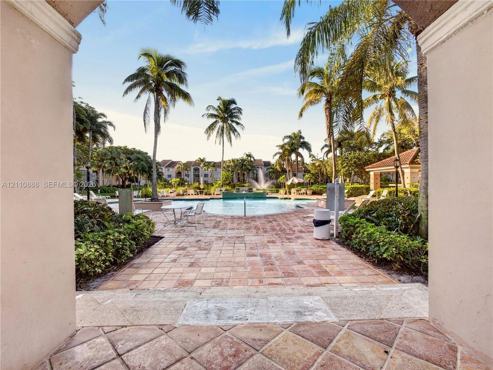 Swimming pool featuring a center fountain, bordered by a paved stone deck, mature palm trees, and landscaped shrubbery
