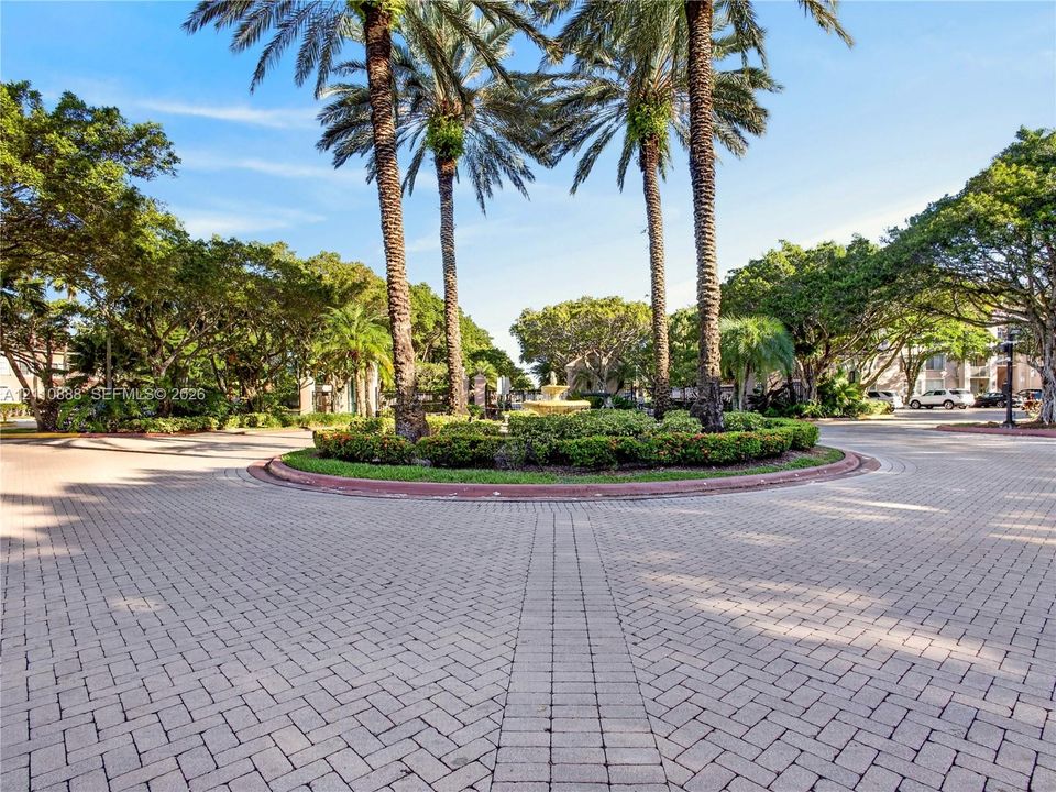 Paver brick circular drive featuring a central garden island, mature palm trees, a tiered stone fountain, and sculpted shrubbery