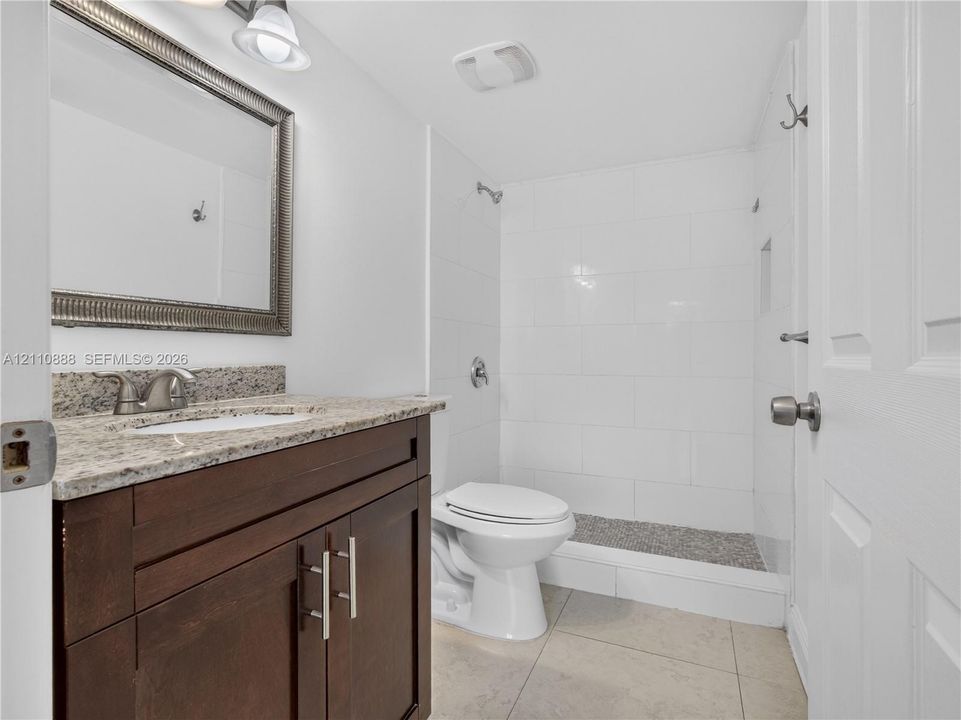 Bathroom featuring a dark wood-finish vanity, stone-patterned countertop with an integrated sink, rectangular-framed wall mirror, and a walk-in shower with white wall tile and stone-texture flooring