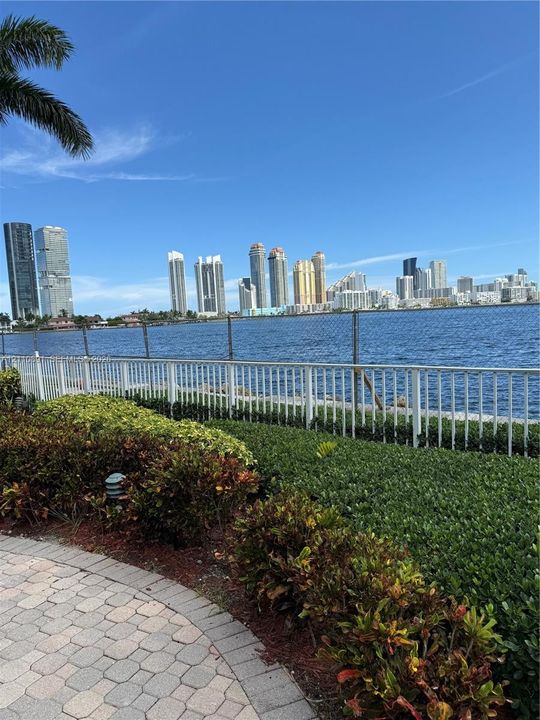 Waterfront property featuring a paved stone walkway, landscaped shrubbery, a white perimeter fence, and a skyline backdrop