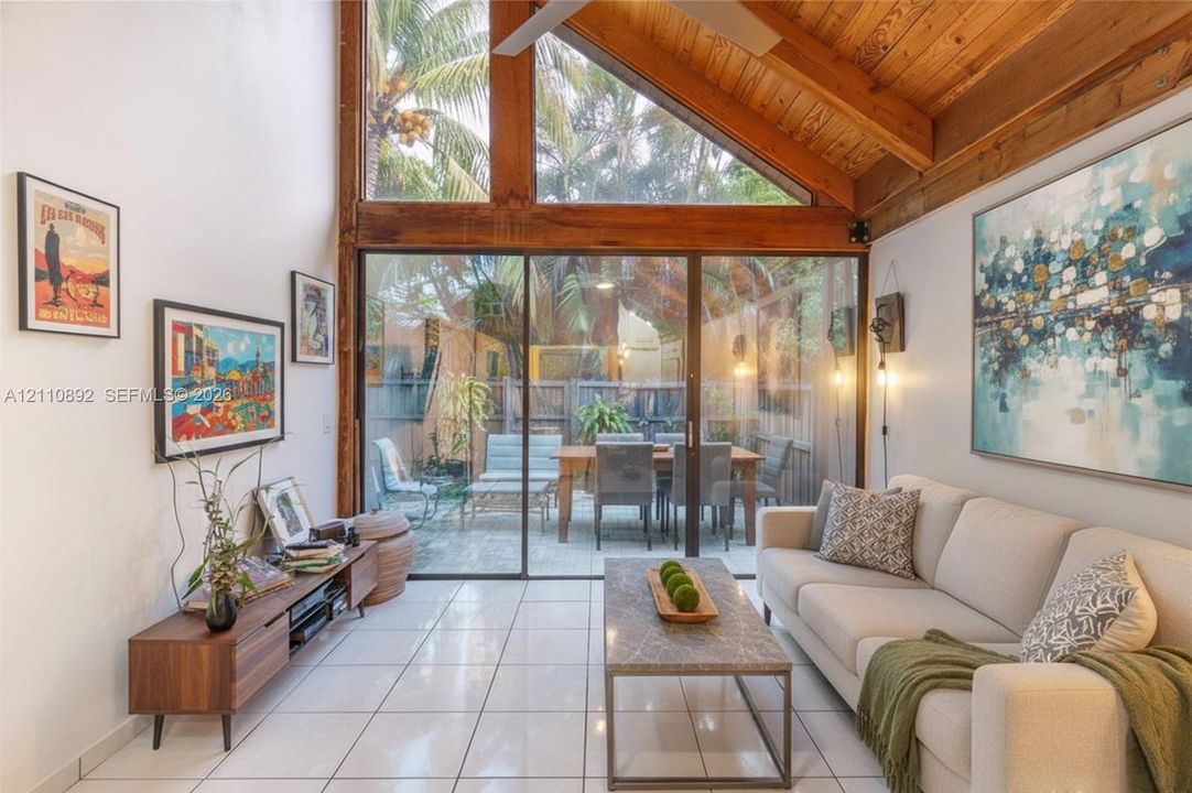 Vaulted ceiling with exposed wood beams, full-height windows, floor-to-ceiling glass sliding doors, and light-toned tile flooring