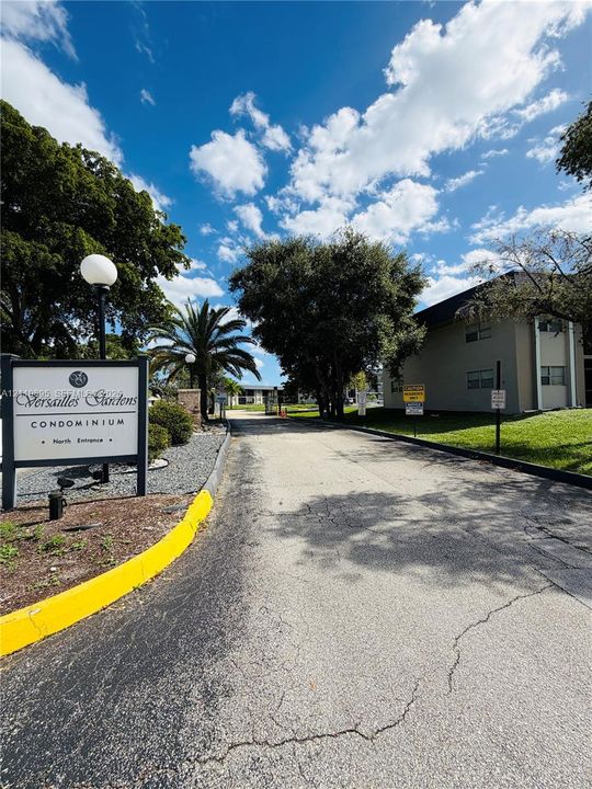 Entrance to Versailles Gardens Condominium featuring mature shade trees, established landscaping, and paved driveway