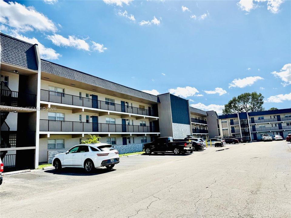 Multi-story exterior featuring a shingled roof, neutral-toned facade, metal railings, and on-site surface parking