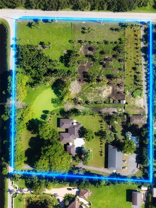 Aerial perspective of a multi-structure property featuring expansive grounds, mature trees, a natural pond, and a paved driveway