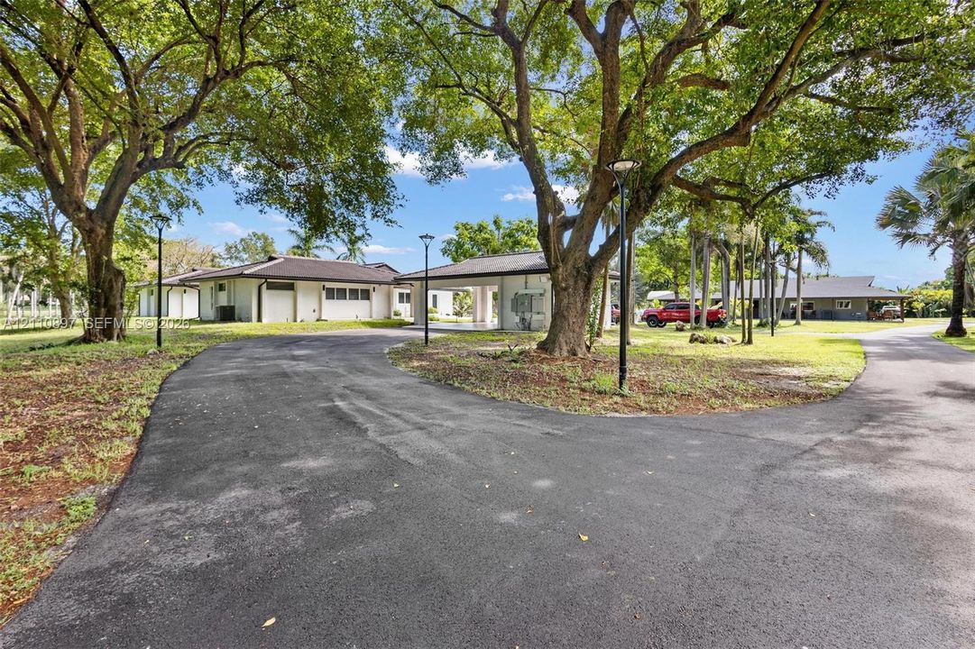 Asphalt circular driveway featuring mature tree canopy, covered porte-cochère, and pole-mounted exterior lighting
