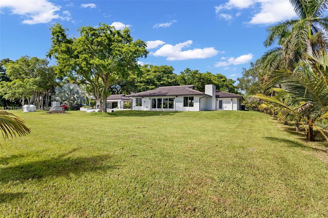 Single-story residence featuring a hipped tile roof, white stucco exterior, expansive lawn, mature specimen trees, and palm landscaping
