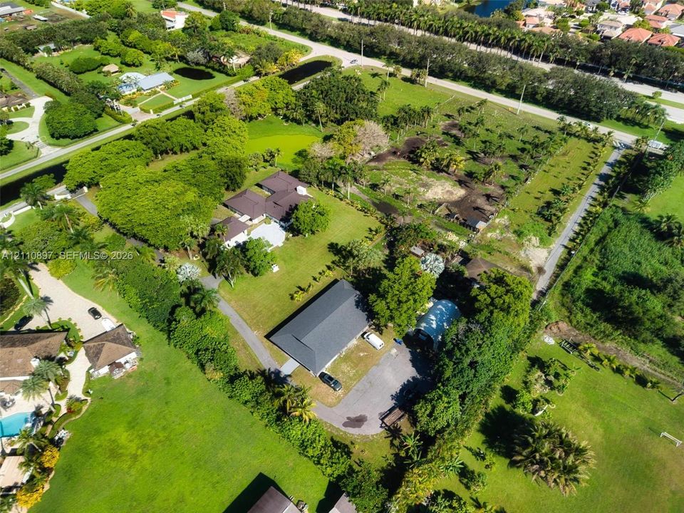 Aerial perspective of a sprawling property featuring extensive open green space, mature canopy trees, asphalt driveways, and multiple residential structures with dark-toned shingled roofing