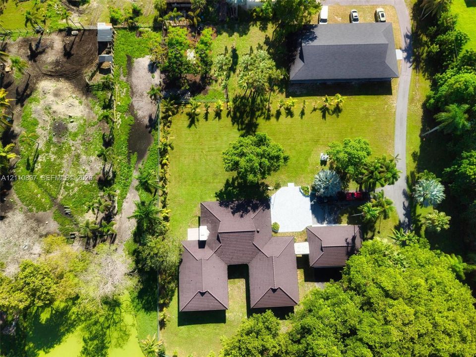 Aerial perspective featuring a sprawling lot with mature trees, a detached multi-vehicle structure, a paved driveway, and a residence with a multi-hipped roofline