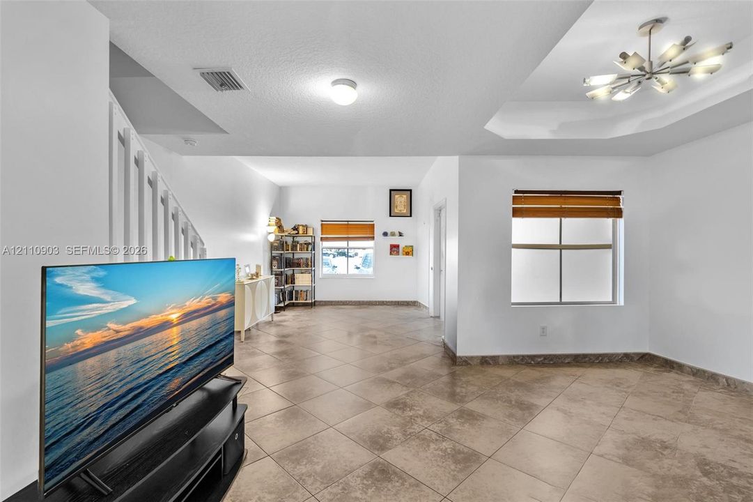 Large-format tile flooring, recessed tray ceiling, multi-bulb modern chandelier, and dual windows with fitted wood-tone blinds