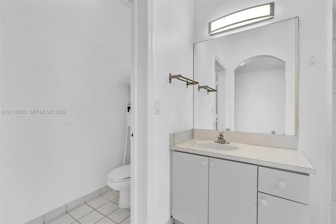 Bathroom vanity featuring light-toned cabinetry, an integrated sink, a full-width mirror, and a wall-mounted light fixture, paired with tile flooring and a standard-height toilet