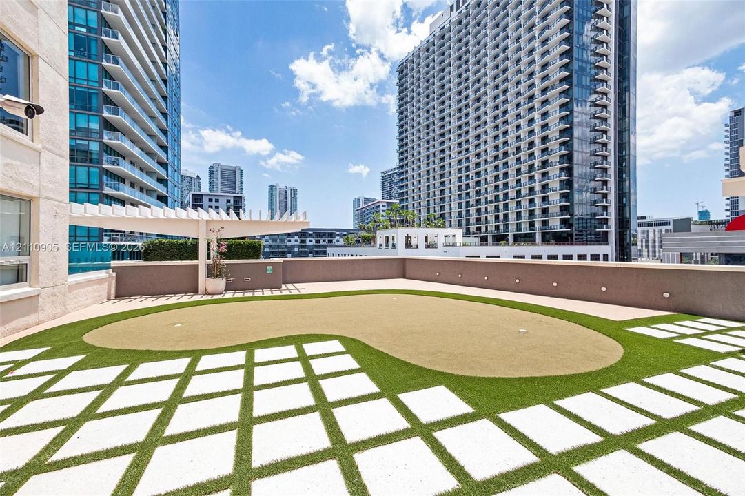 Rooftop terrace featuring a synthetic turf putting green, geometric paver hardscaping, built-in concrete wall enclosure, and a white architectural pergola