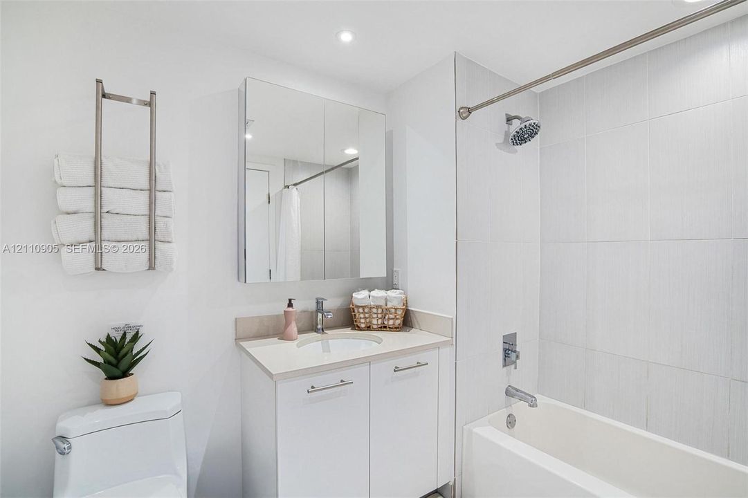 White vanity with solid-surface countertop and integrated basin, mirrored medicine cabinet, wall-mounted towel rack, and full-height tiled shower surround with bathtub