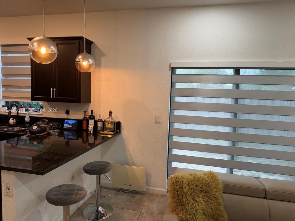 Dark cabinetry with polished stone countertops, dual glass pendant light fixtures, and dual-layered cellular window shades