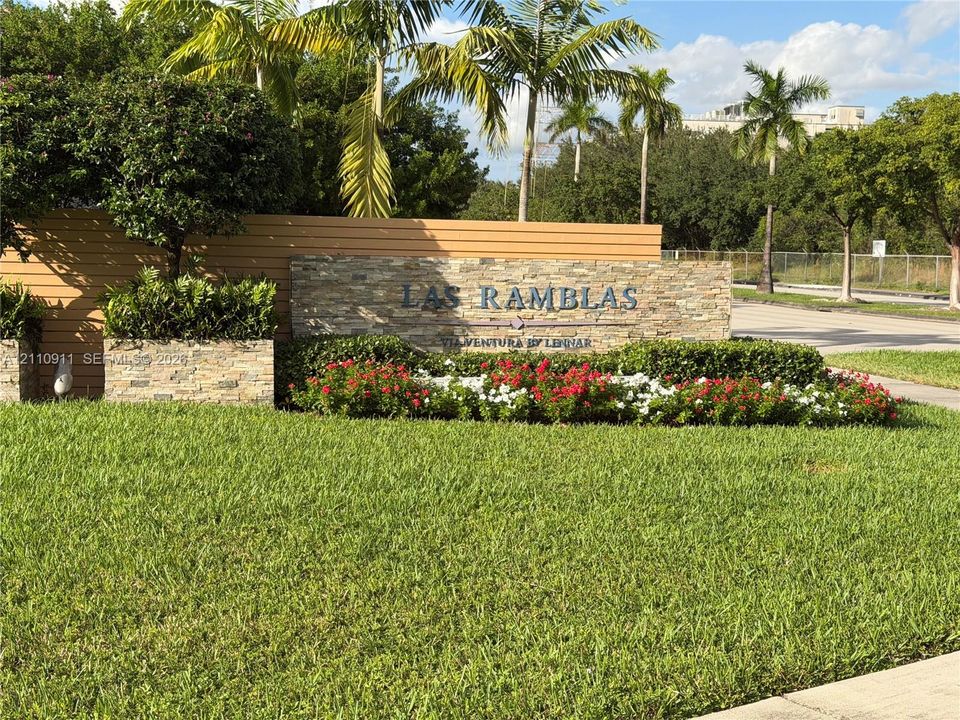 Community entrance signage featuring stacked stone masonry, horizontal wood-panel accenting, and landscaped garden beds with mature palms