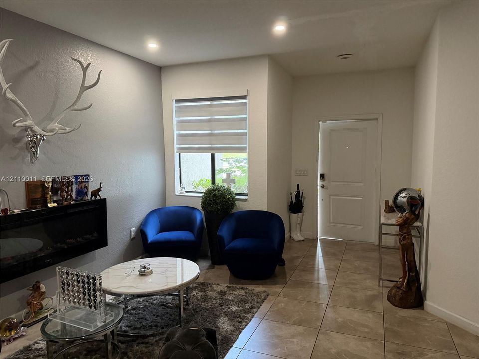 Neutral-toned walls featuring a recessed electric fireplace and light-colored tile flooring throughout, accented by recessed lighting and a window fitted with dual-layer cellular shades