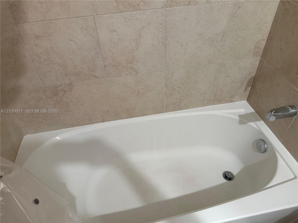 Built-in soaking tub with a white composite finish, accented by metallic fixtures and surrounded by neutral-toned square wall tile