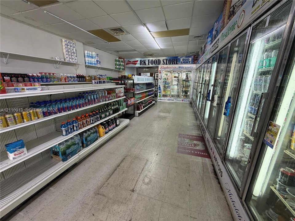 Retail space featuring multi-tiered metal shelving, a wall-length row of reach-in glass display coolers, drop ceiling tiles, and recessed fluorescent lighting