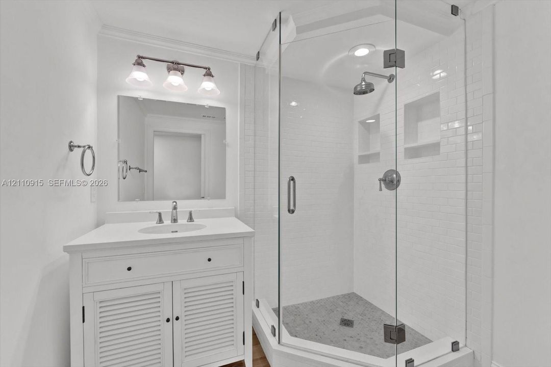 White vanity with louvered cabinet doors and countertop-integrated basin, glass-enclosed shower with subway tile surround, recessed wall niches, and brushed-nickel hardware