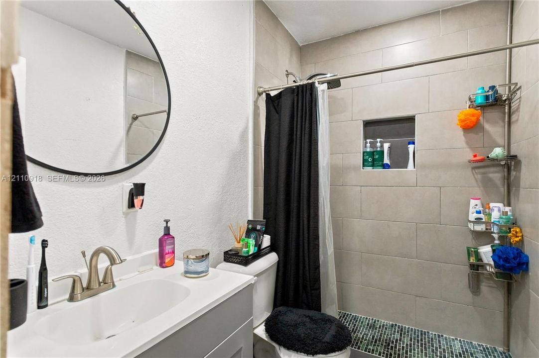 Bathroom featuring a vanity with a white countertop and integrated sink, matte-finish faucet, circular wall mirror, and a shower area with tiled walls and a recessed niche