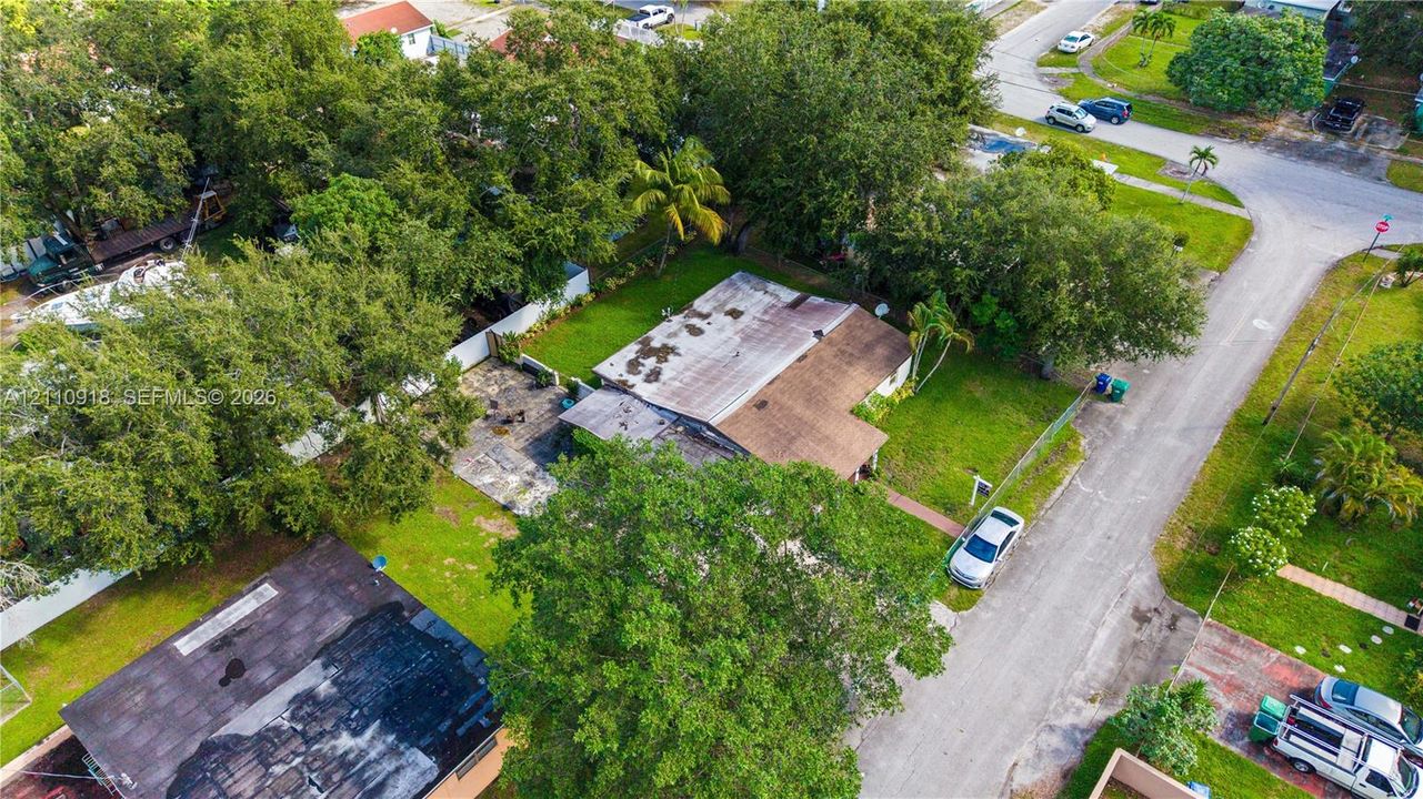 Aerial perspective of a corner lot featuring established mature trees, a designated concrete patio, and a shingled roof exterior