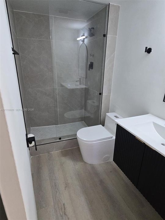 Bathroom featuring a glass-enclosed shower with large-format tile surround, black plumbing fixtures, wood-finish flooring, and a modern vanity with a fluted cabinet base and white countertop