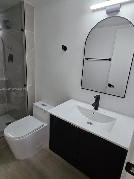 Bathroom featuring a black fluted-front vanity with a white countertop, an arched wall mirror with a black frame, a walk-in shower with large-format tile surround, matte black hardware, and a one-piece elongated toilet