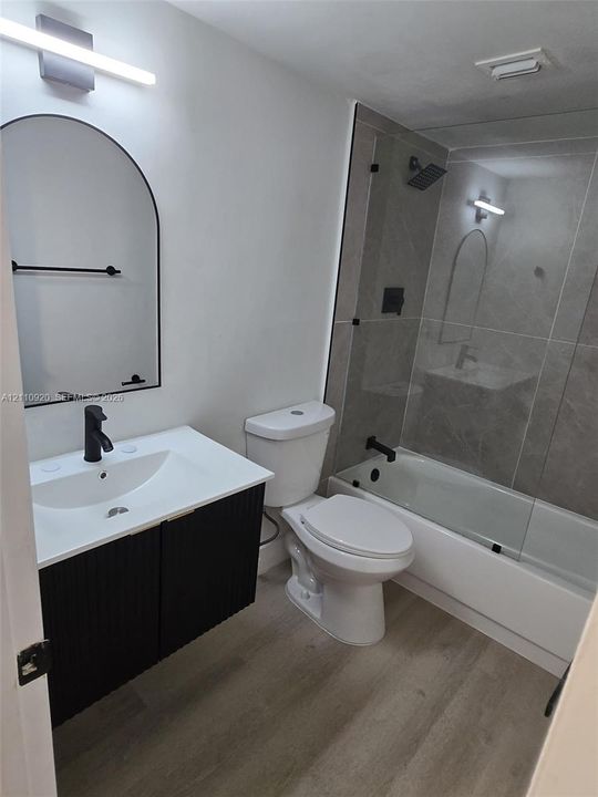 Bathroom featuring a black fluted vanity with a white countertop, an arched mirror, wood-finish flooring, and a shower-tub combination with large-format tile surround and matte black fixtures