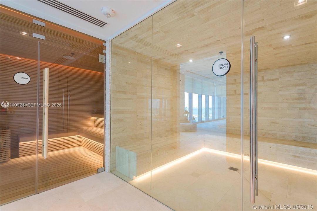 Wellness area featuring a glass-enclosed wood sauna with tiered bench seating; a separate glass-enclosed steam room with stone tile walls, and recessed under-bench lighting